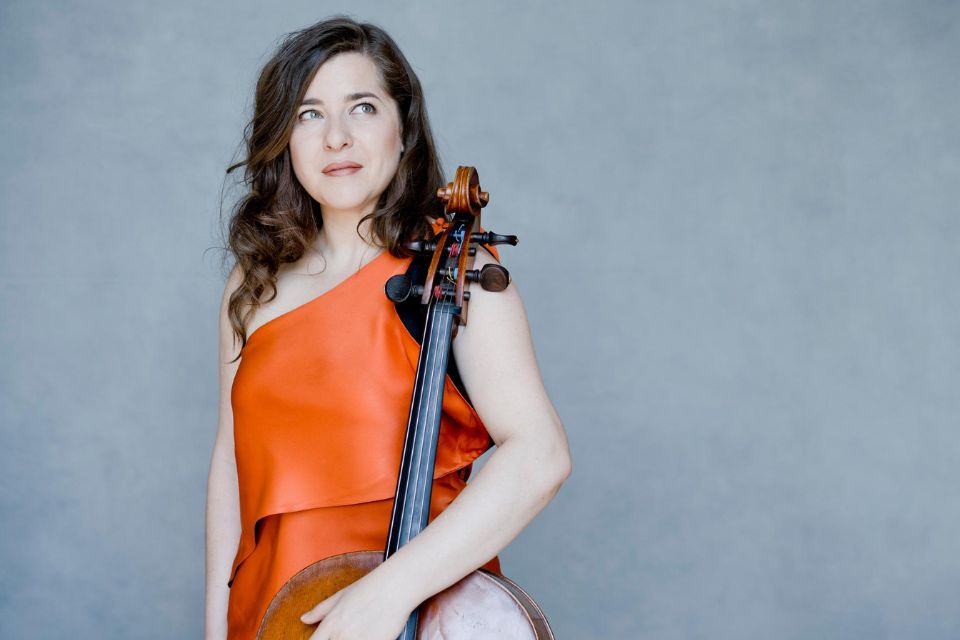 Alisa Weilerstein wears a bright orange dress against a pastel blue background, holding her cello and looking to the side